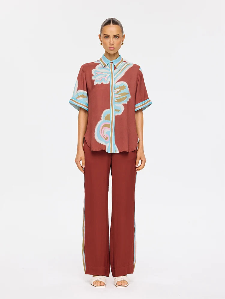 A model stands against a plain white studio background wearing a matching rust-red two-piece set consisting of a short-sleeve button-up shirt with abstract light blue, white, and gold patterns, and wide-leg trousers with light blue side stripes.