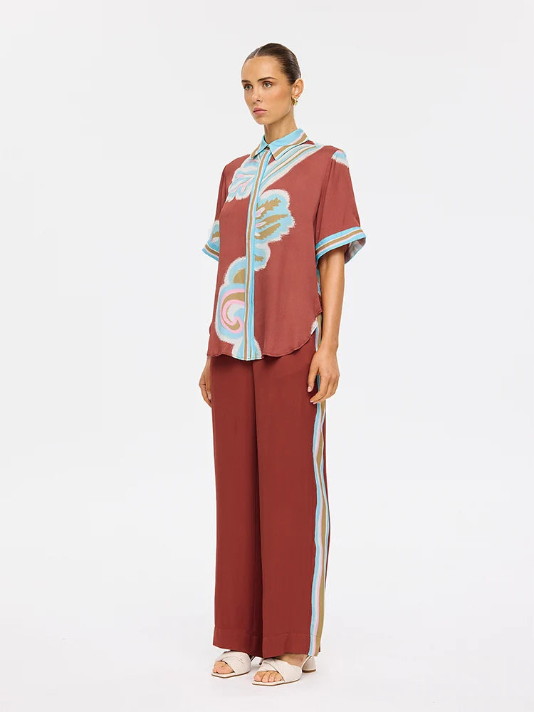 A model stands against a plain white studio background wearing a matching rust-red two-piece outfit with a short-sleeve button-front top featuring light blue and cream abstract print details, and wide-leg pants with light blue side stripes.