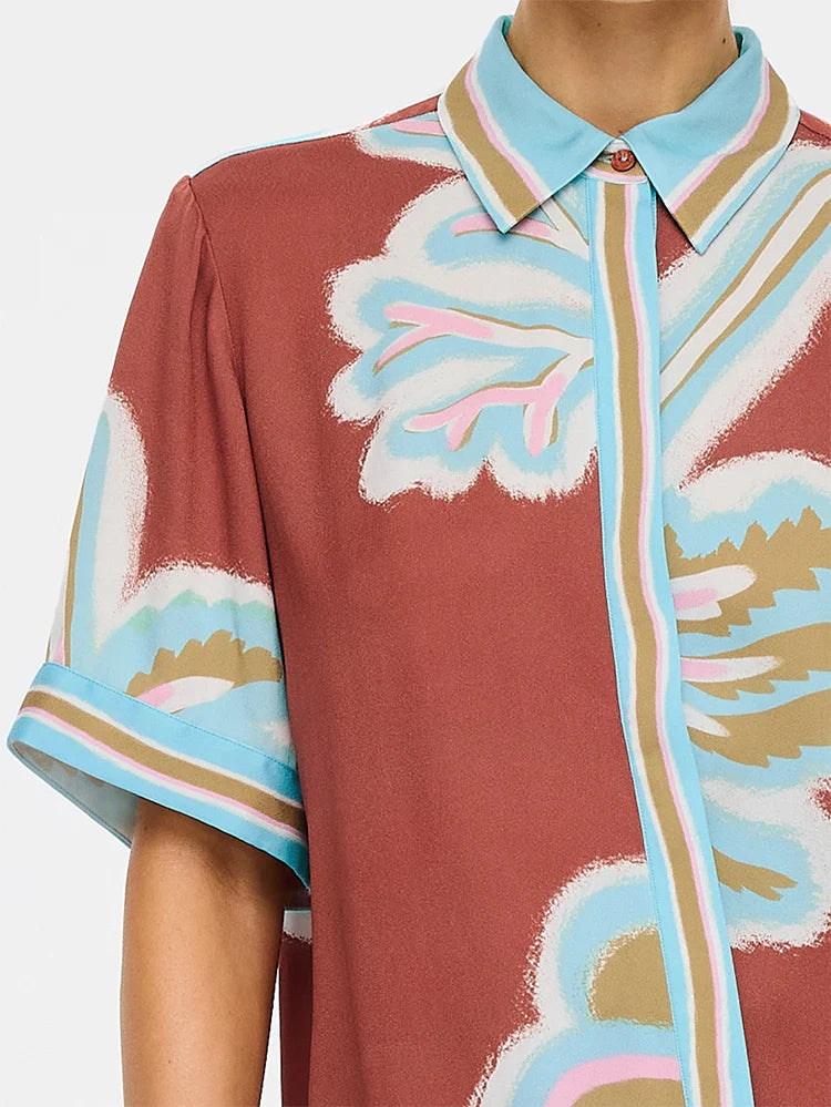 A close-up of a short-sleeve button-front shirt with a bold abstract floral print. The shirt features a terracotta brown base with light blue, white, pink, and tan patterns, shown on a model against a plain white background.