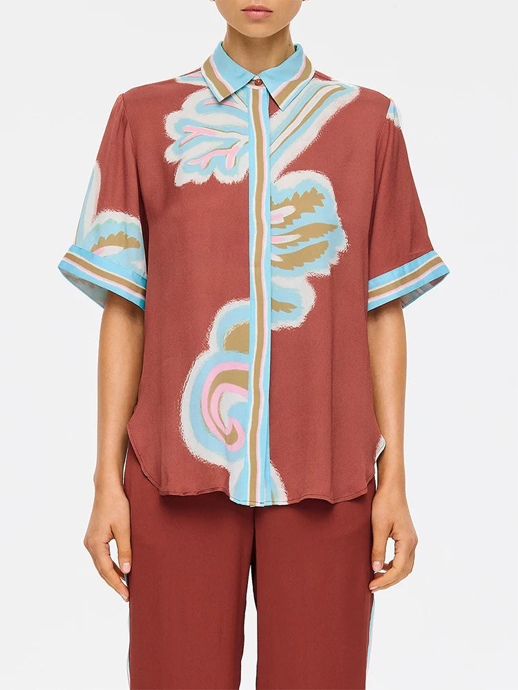 A model wearing a short-sleeve button-up shirt with a rust-brown base and a colorful abstract print in light blue, beige, and white, paired with matching rust-brown trousers against a light gray background.