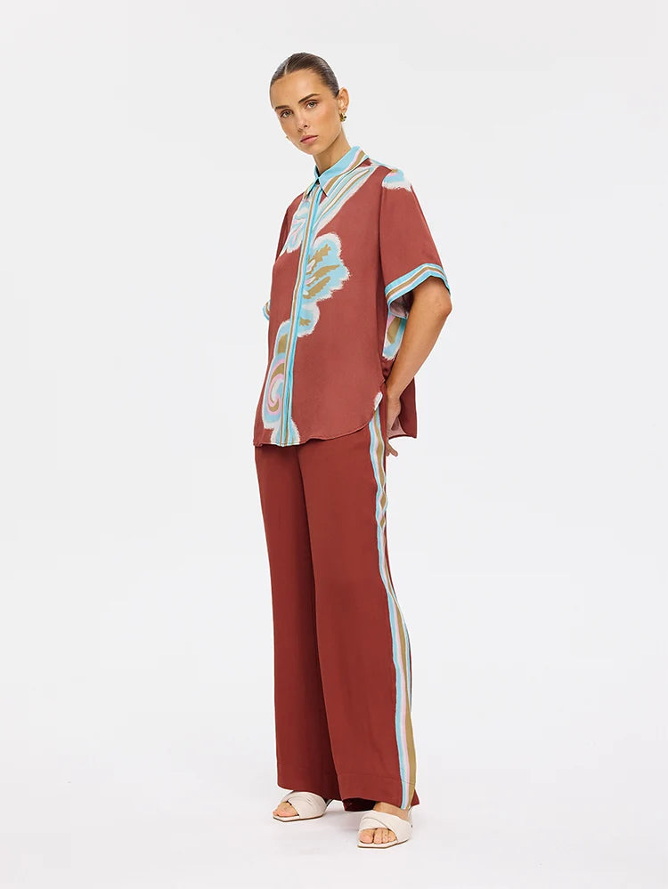 A model stands against a plain white studio background wearing a matching rust-red two-piece set with a short-sleeve button-front top and wide-leg pants. The set features light blue and beige abstract stripe and wave patterns along the collar, placket, sleeve cuffs, and down the outer side of the pants.