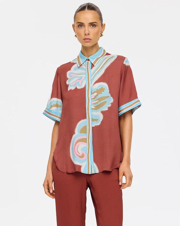 A model wearing a rust-brown short-sleeve button-up shirt with a light blue, pink, and beige abstract floral print, paired with matching rust-brown trousers, against a light gray background.