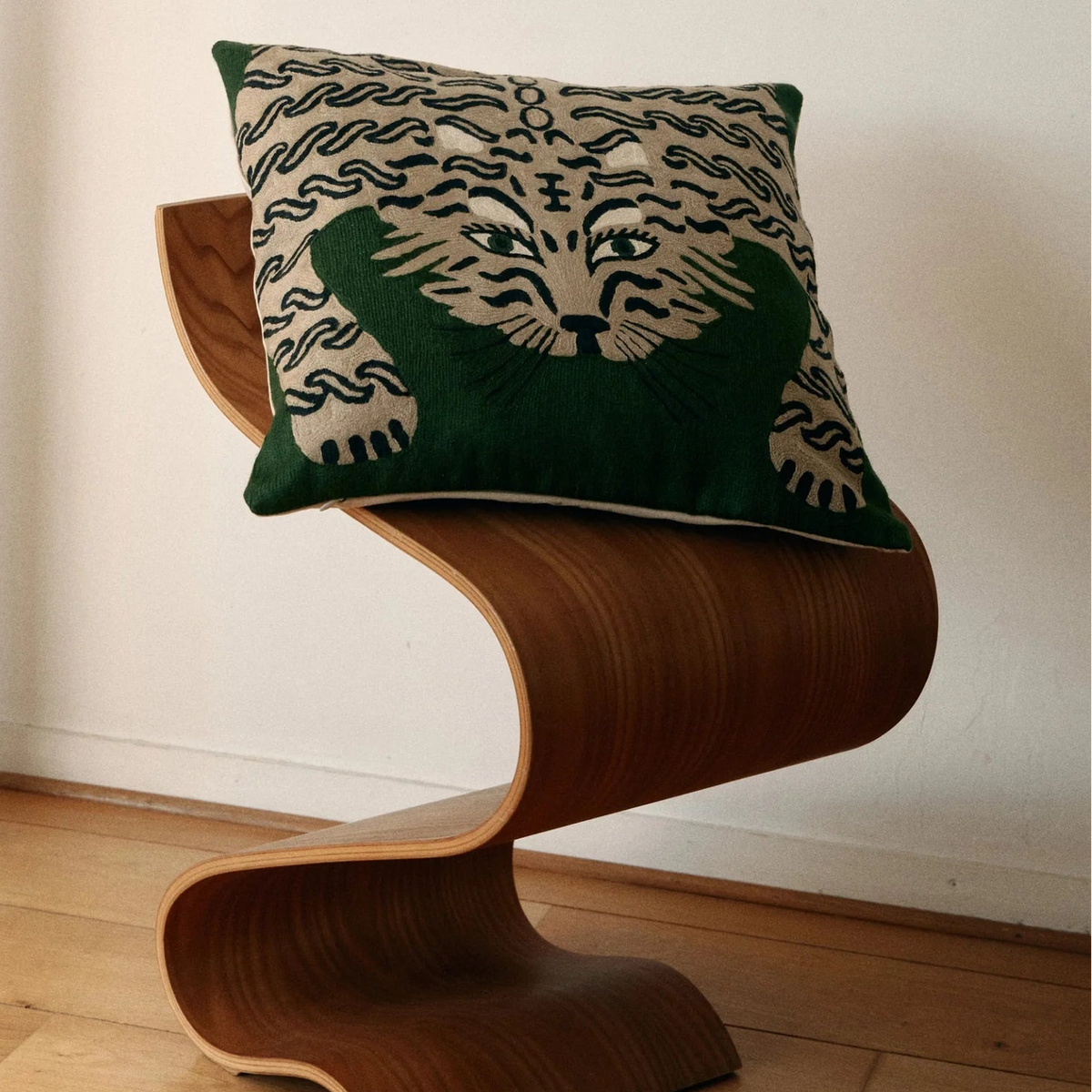 A decorative throw pillow with a green and beige patterned design, resting on a sculptural curved wooden stool in a minimalist room with light wood floors and a white wall.