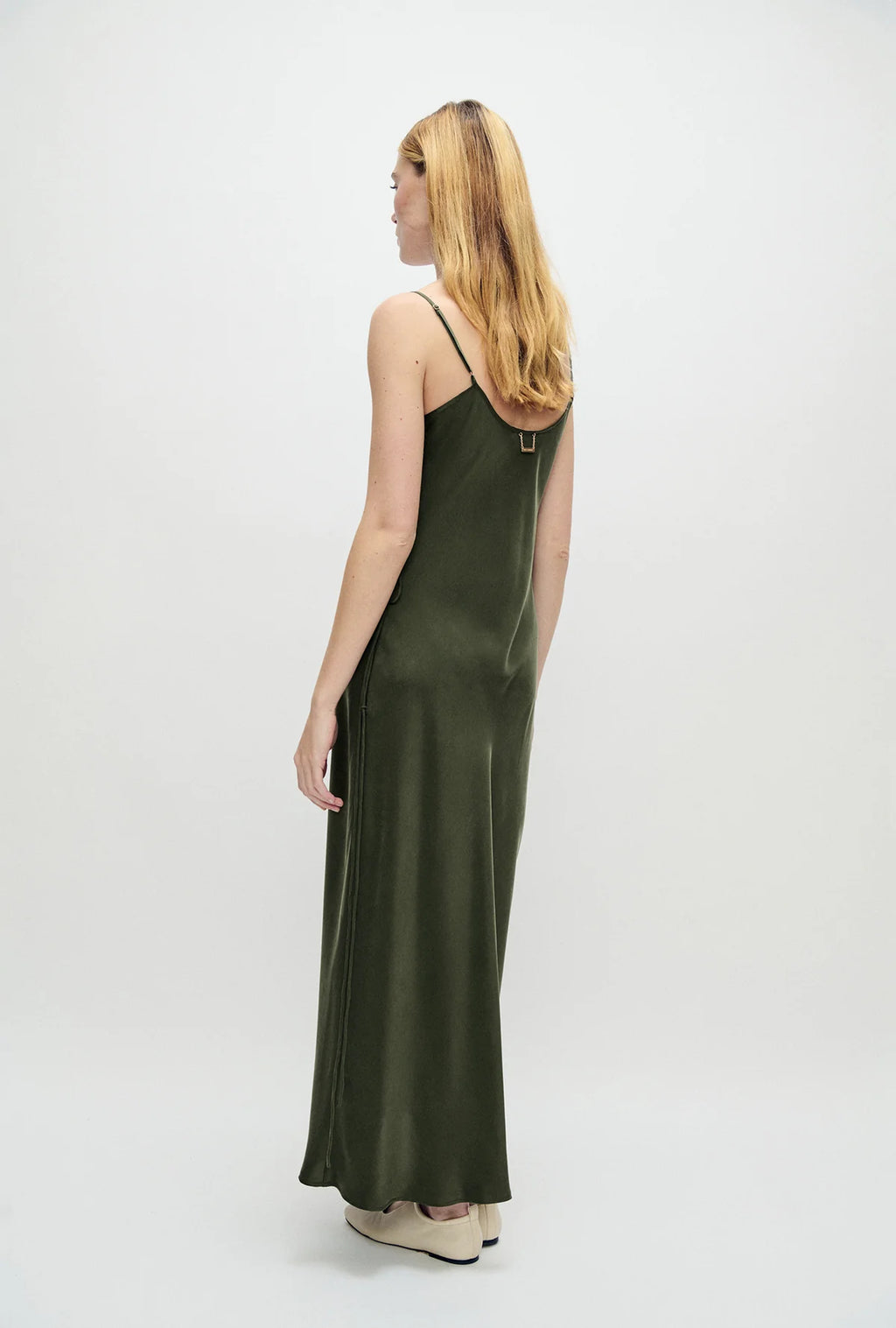 A woman with blonde hair is shown from the back wearing a dark olive green satin slip dress with thin straps and a small gold accent at the back neckline, set against a plain white studio background.