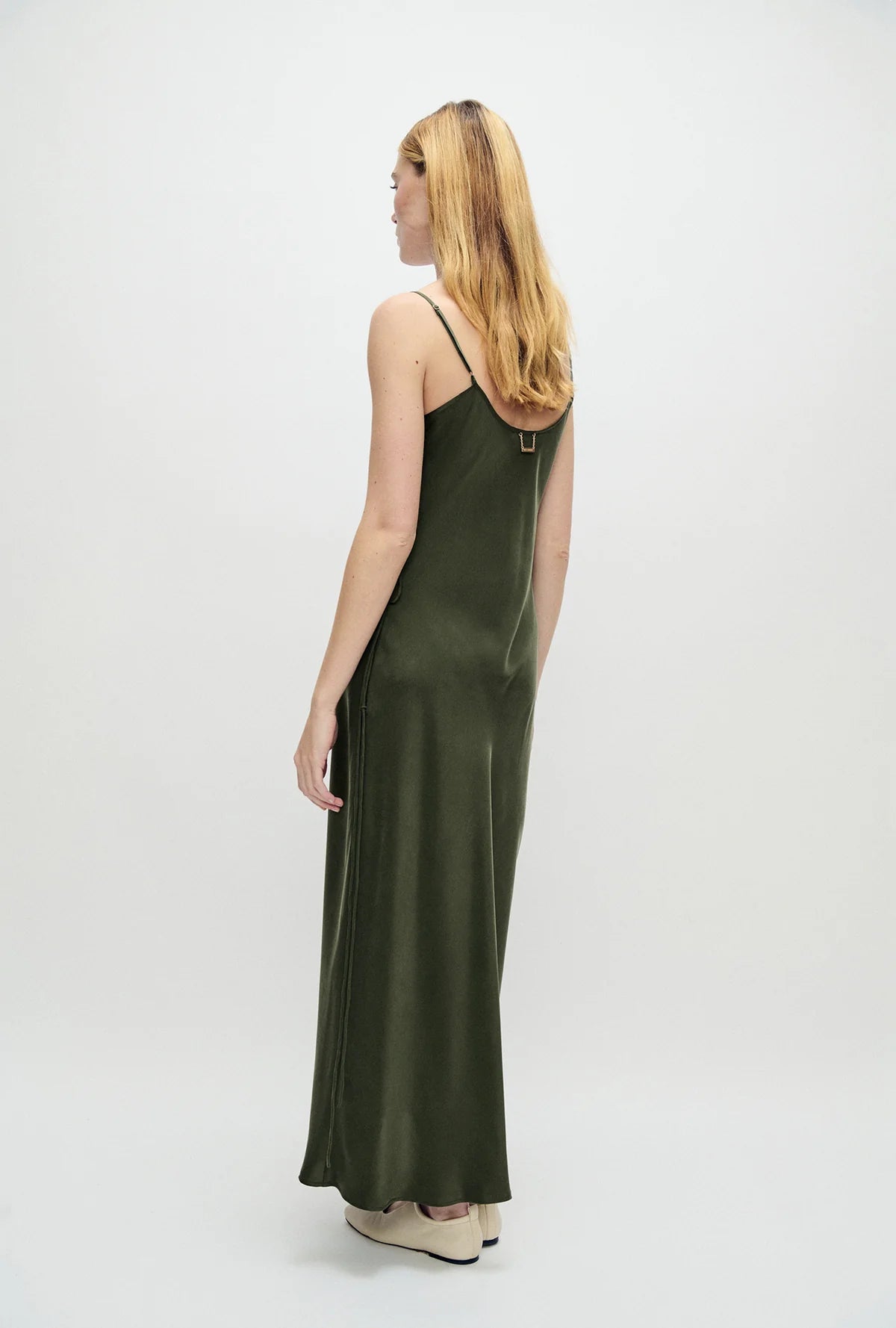 A woman with blonde hair is shown from the back wearing a dark olive green satin slip dress with thin straps and a small gold accent at the back neckline, set against a plain white studio background.