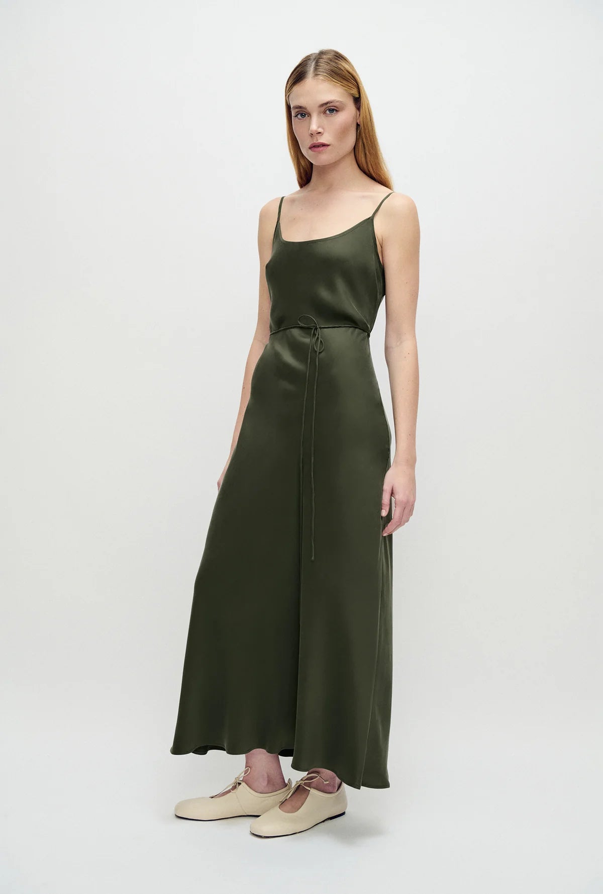 A woman modeling a dark olive green satin midi dress with spaghetti straps and a drawstring waist, paired with beige flat shoes, against a plain light gray studio background.
