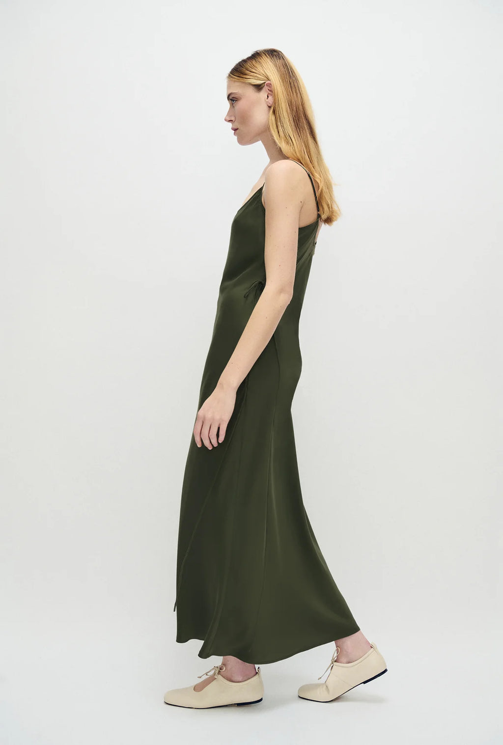 A woman in profile wearing a long olive green satin slip dress and beige flat shoes, standing against a plain light gray studio background.