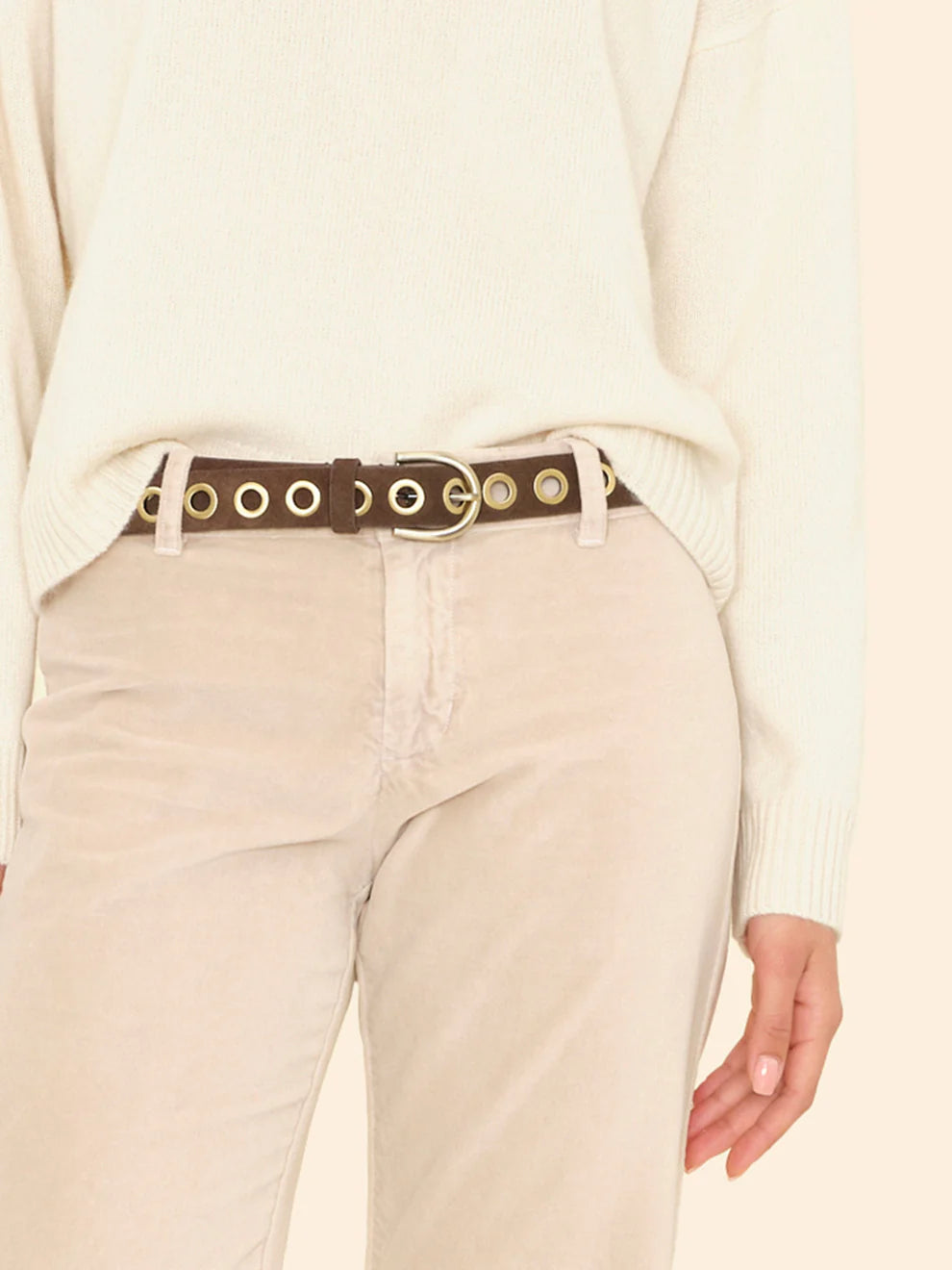 Person wearing a beige sweater and pants with a belt featuring gold accents on a plain background